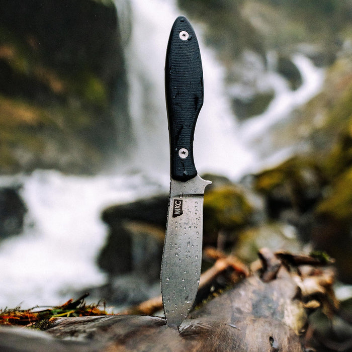 Black JACKSTONE knife standing on a log near a waterfall, showcasing its durable blade and textured handle.