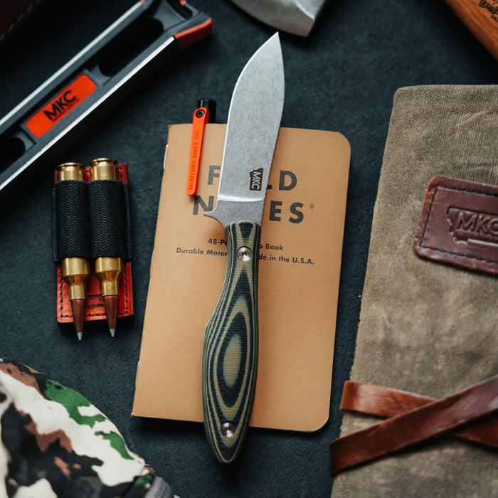 JACKSTONE knife in green and black with a sleek blade, placed on a notebook surrounded by outdoor gear.