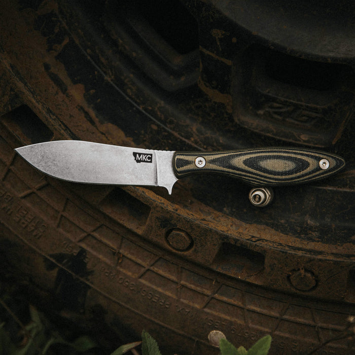 Green and black JACKSTONE knife resting on a muddy tire, showcasing its durable blade and ergonomic handle design.