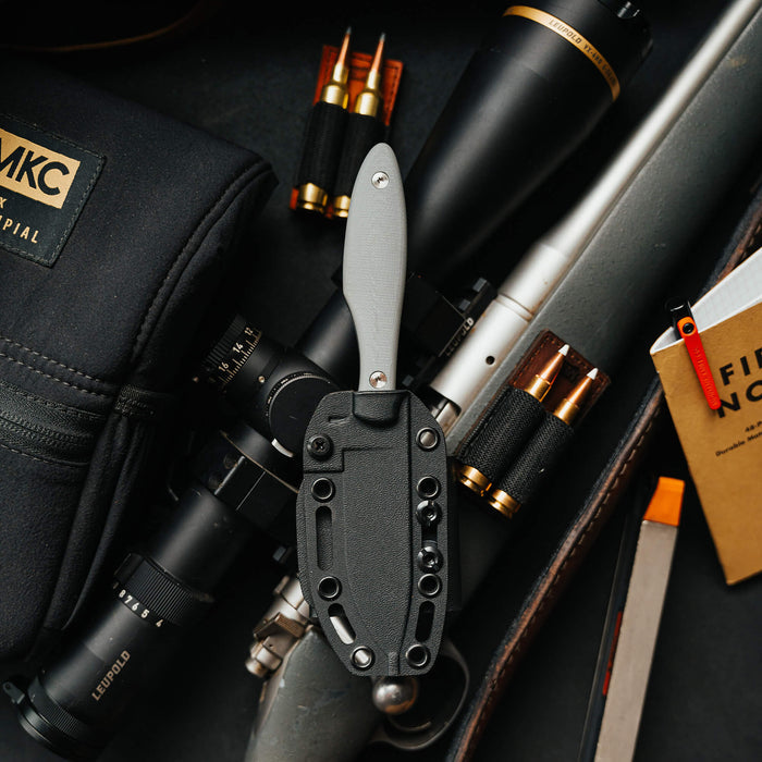 JACKSTONE knife in grey positioned on a dark surface with a rifle scope, ammunition, and a notebook.