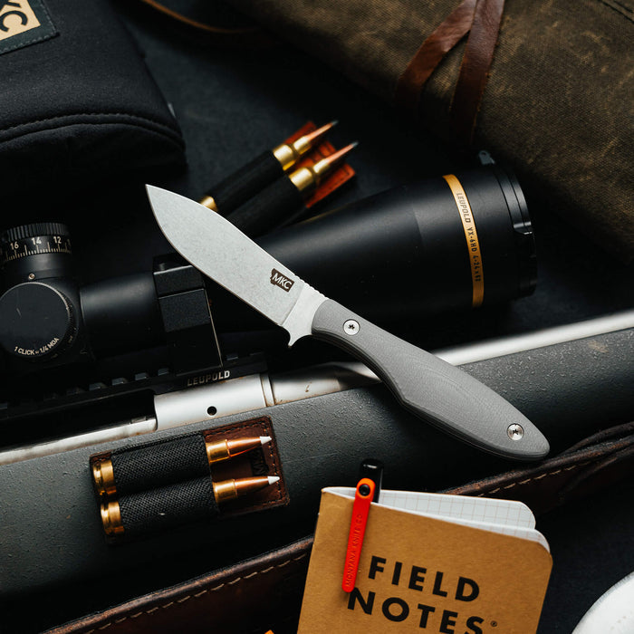 Grey JACKSTONE knife displayed among outdoor gear, including a scope, ammunition, and a notebook, showcasing its rugged design.