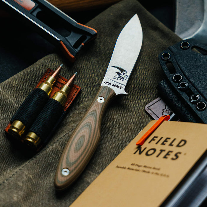 Olive and tan JACKSTONE knife with a textured handle, placed on a rugged surface alongside ammunition and a notebook.
