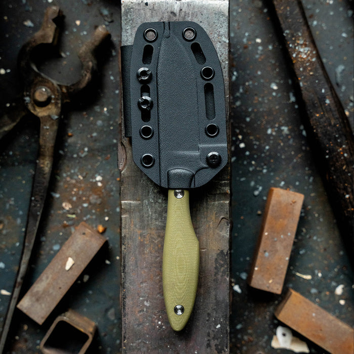 Olive JACKSTONE knife with a textured handle and sheath, displayed on a workbench with tools and materials.