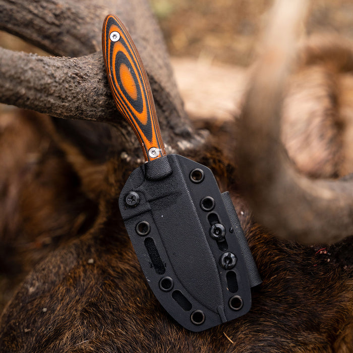 JACKSTONE knife with orange and black handle, secured in a black sheath, resting on a textured surface.