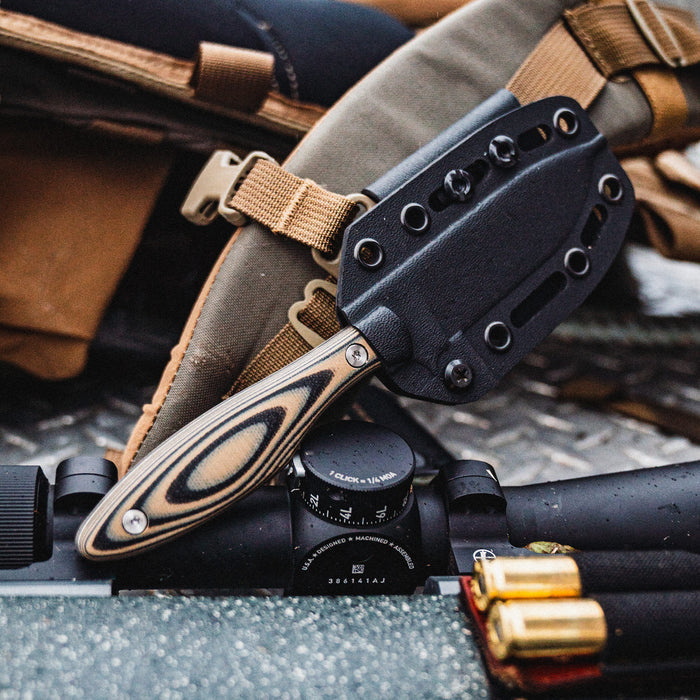 Jackstone knife in tan and black resting on a tactical setup with a scope and ammunition, showcasing its durable design.