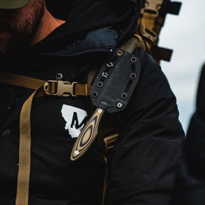JACKSTONE knife in tan and black, secured on a person's shoulder strap, showcasing its rugged design and functionality.