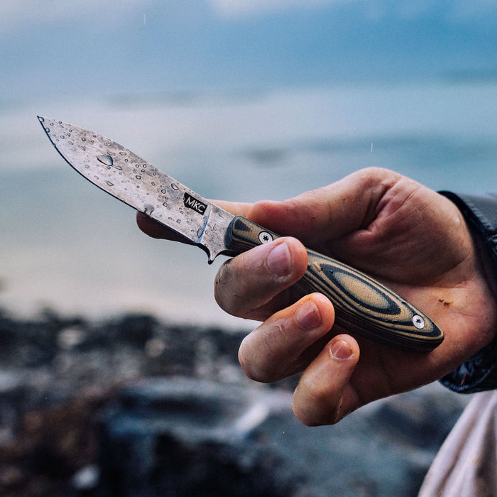 Hand holding the JACKSTONE knife featuring a tan and black handle and a patterned blade, set against a natural outdoor backdrop.