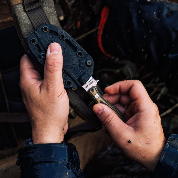 A person securing the JACKSTONE knife in a tan and black sheath, showcasing its rugged design and functionality.