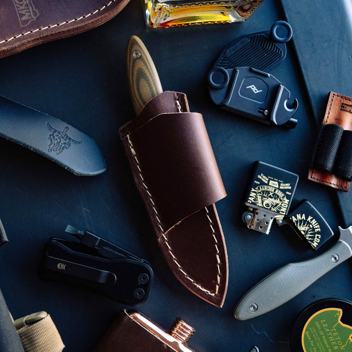 JACKSTONE leather sheath designed for horizontal belt carry, surrounded by various knives and tools on a dark surface.
