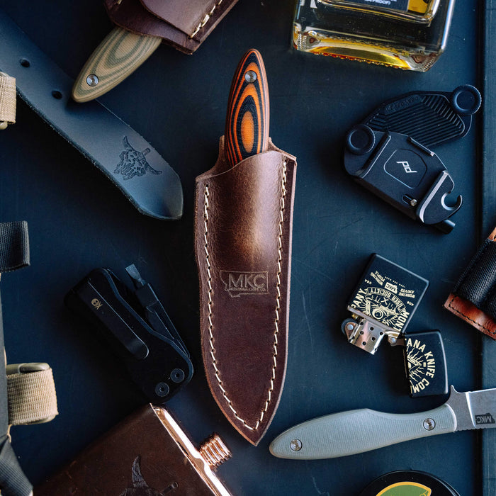 Vertical leather sheath for knives, featuring a sleek design and MKC branding, surrounded by various tools and accessories.
