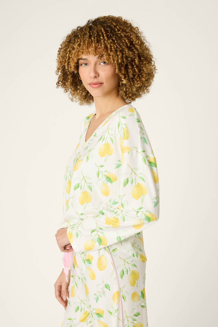 A person with curly hair stands sideways, looking at the camera against a plain light background, wearing a lemon-themed top.