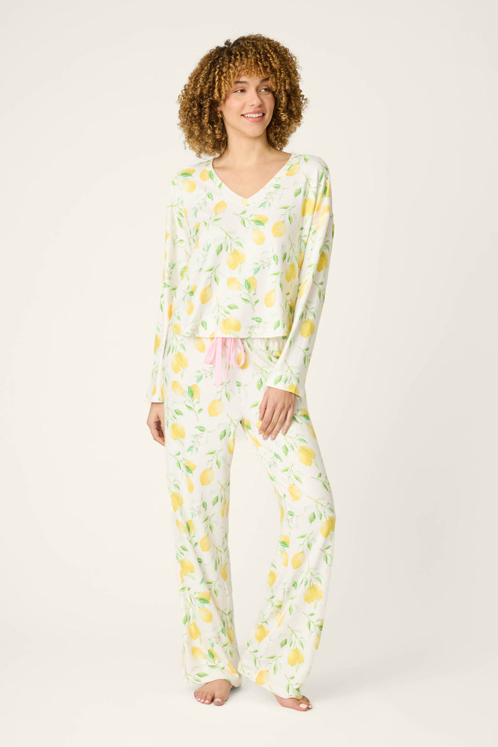 A person with curly hair wears pajama pants with a pink drawstring and a matching long-sleeve V-neck top, barefoot on a plain background.