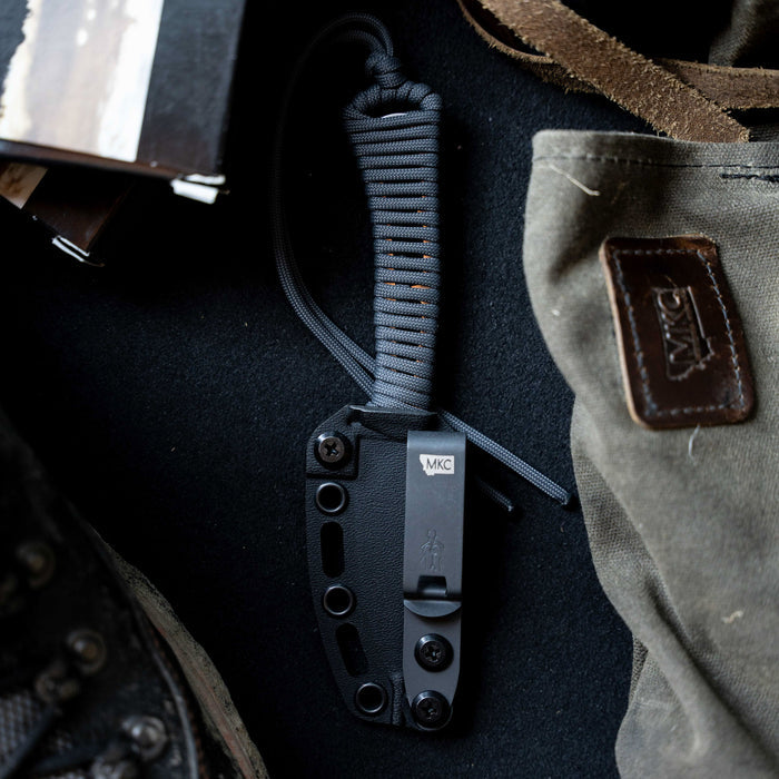 MAGNACUT MINI SPEEDGOAT 2.0 knife in grey with a paracord handle, resting on a textured surface with gear.
