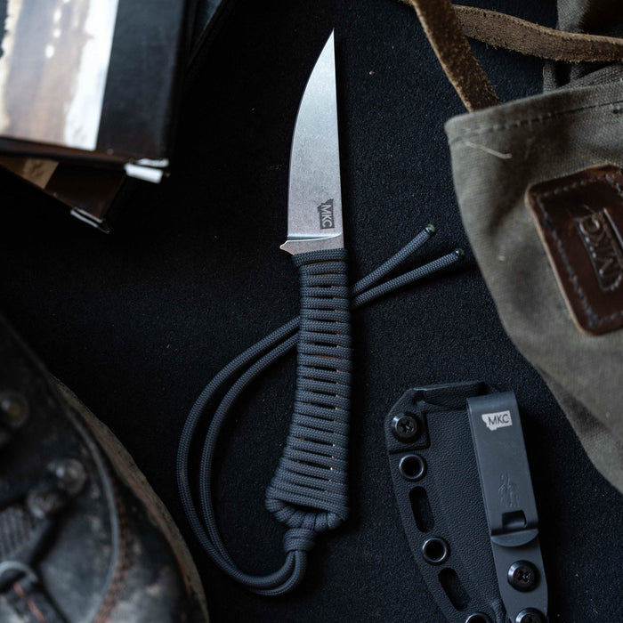 Grey MAGNACUT MINI SPEEDGOAT 2.0 knife with paracord handle, surrounded by outdoor gear and tools.