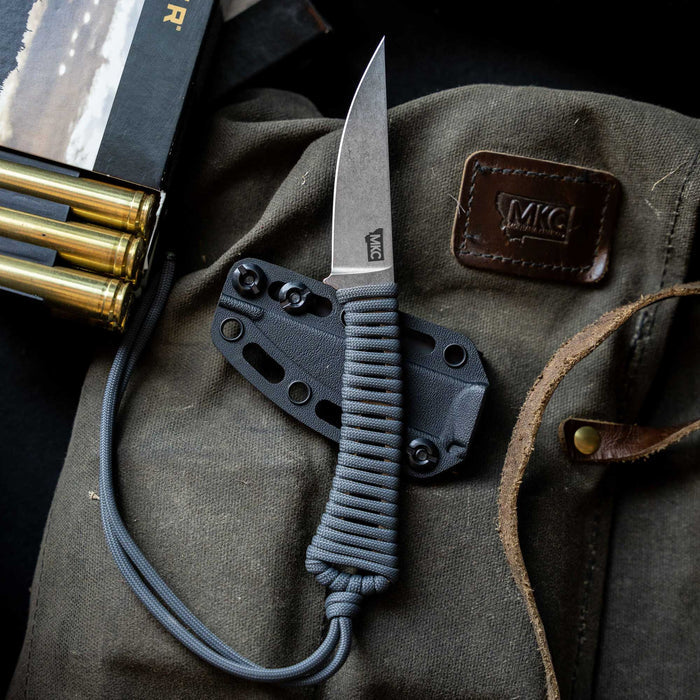 MAGNACUT MINI SPEEDGOAT 2.0 knife in grey with a paracord handle, displayed on a rugged canvas background with ammunition.
