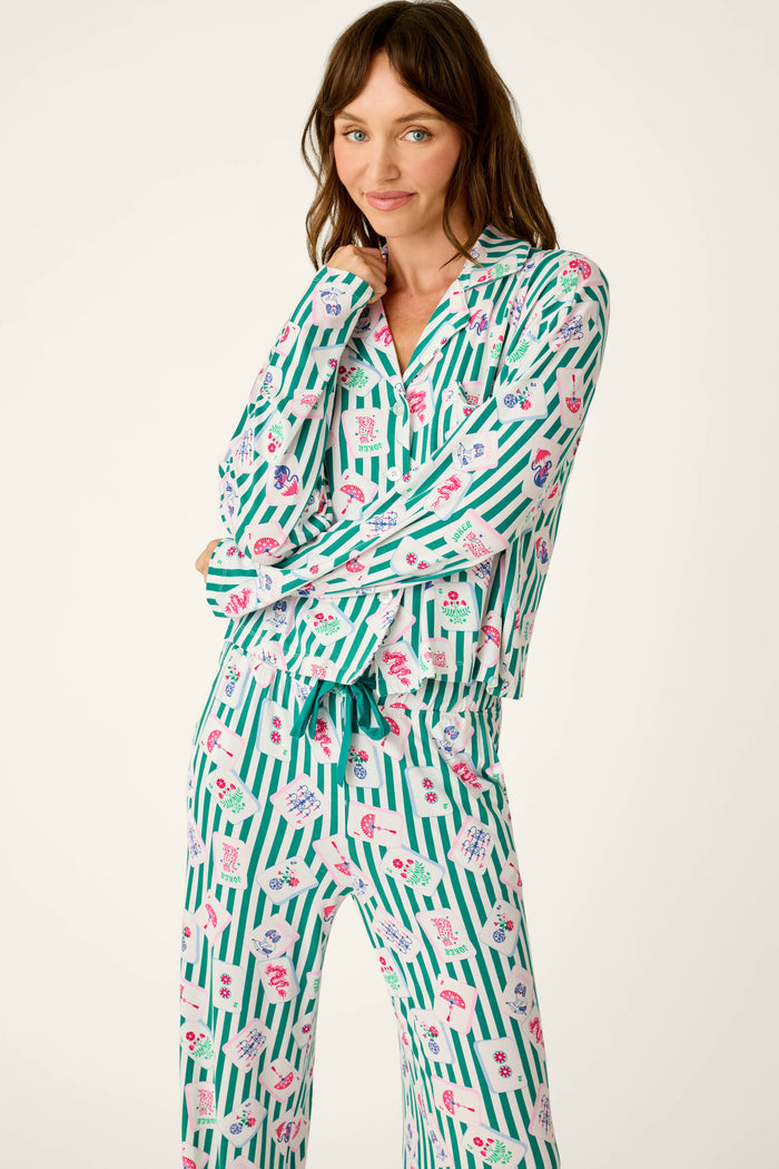 A woman wearing the P.J. Salvage Mahjong Queen Dreams Mahjong Print Long PJ Set, featuring green and white stripes with colorful floral patterns, stands with one arm crossed and smiles at the camera.