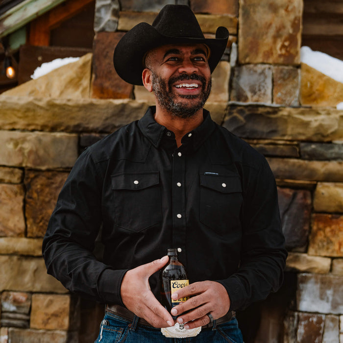 Man wearing a black pearl snap long sleeve shirt, smiling while holding a drink, set against a stone wall backdrop.