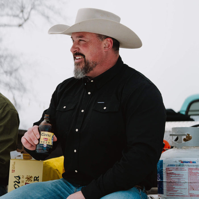 Man wearing a black pearl snap long sleeve shirt and cowboy hat, holding a beverage while sitting outdoors.