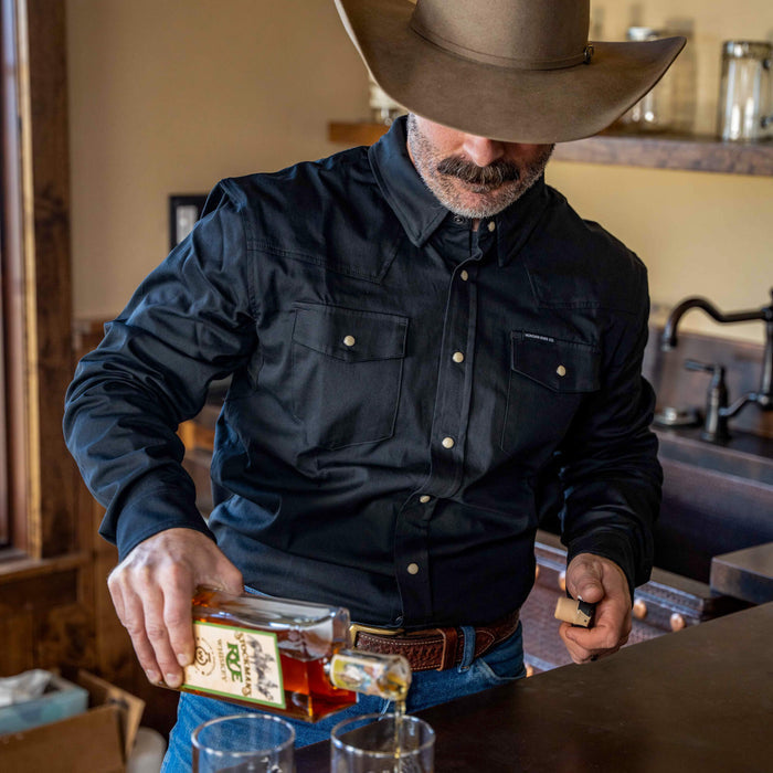A man in a black long sleeve pearl snap shirt pours whiskey into glasses at a rustic bar.