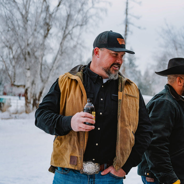 Man wearing a black pearl snap long sleeve shirt under a brown vest, holding a drink outdoors in a snowy setting.