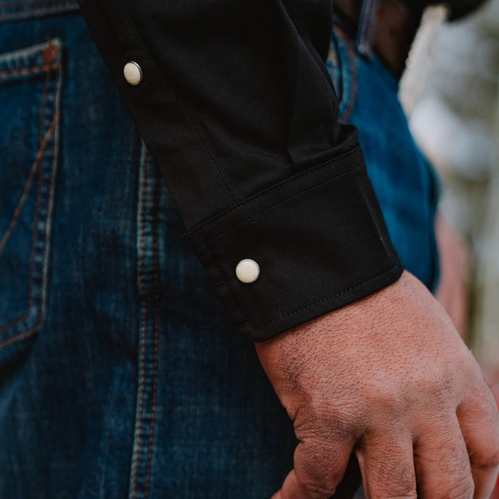 Black long sleeve shirt featuring pearl snap buttons on the cuffs, paired with denim jeans. Ideal for casual or outdoor wear.