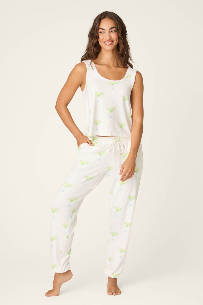 A woman stands barefoot, smiling, in P.J. Salvage's Margarita Mama Spicy Sips Tank Top—a sleeveless white pajama tank with a light margarita print and loose fit. The background is plain and light-colored, evoking relaxing vacation vibes.