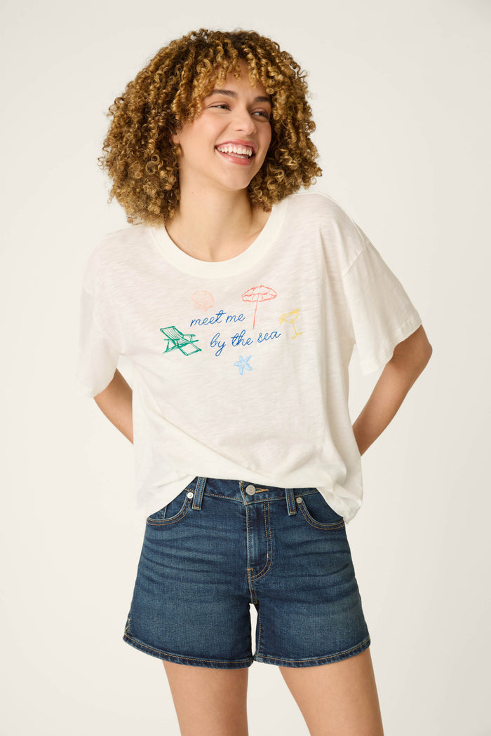 A curly-haired person wears a graphic T-shirt with colorful beach line art and text, blue denim shorts, smiling against a light background.