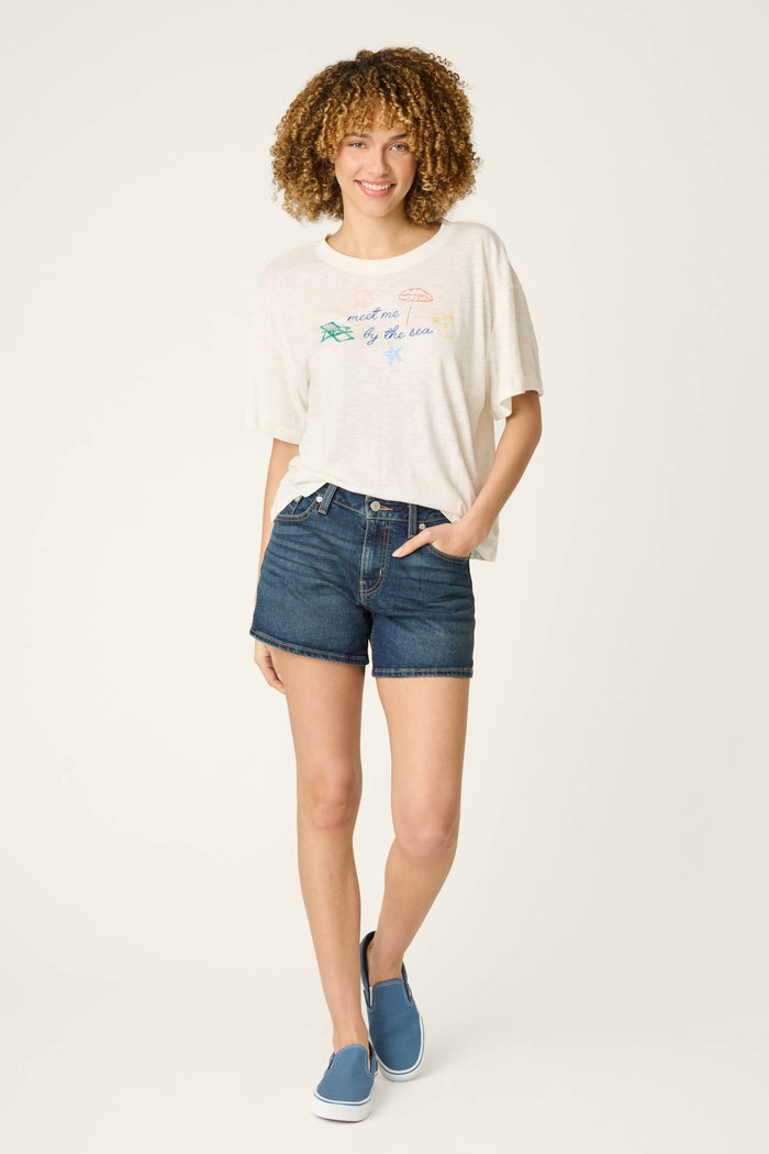 A person with curly hair wears a graphic T-shirt, denim shorts, and blue slip-ons, standing against a plain light background.