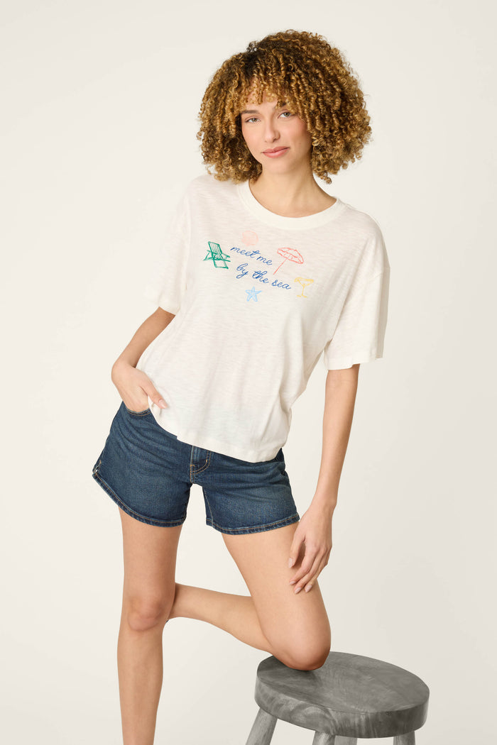 A woman with curly hair wears a graphic print T-shirt and blue denim shorts, standing with one hand in her pocket, one knee on a gray stool.