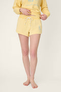 A person stands barefoot adjusting the waistband of light yellow shorts, paired with a matching sweatshirt against a plain, light background.