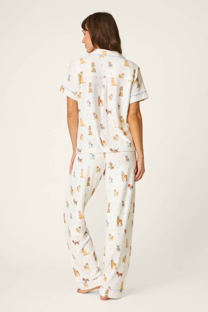 A woman stands barefoot facing away, wearing the P.J. Salvage Meet Me in Pawris Dog Print 3-Piece PJ Set. The set features white pajamas with a colorful dog print, including a short-sleeve top and long pants, and she is posed against a plain, light background.