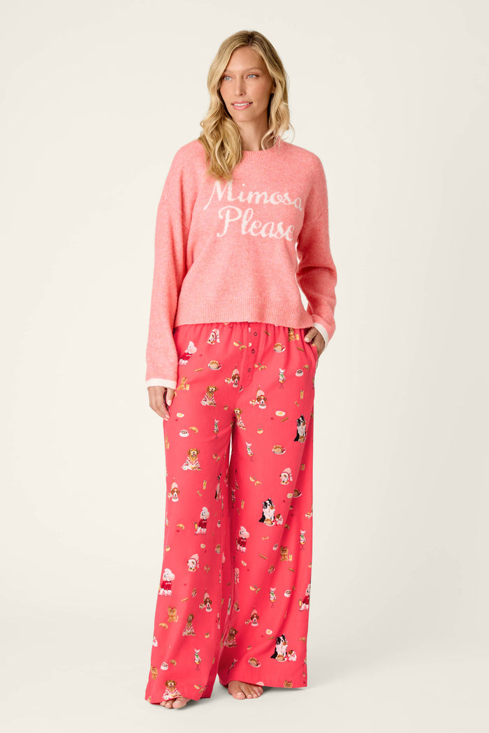 A woman stands and smiles against a plain background, wearing the P.J. Salvage Mimosa Please Snuggle Sweater in pink with “Mimosa Please” text, paired with matching wide-leg pajama pants featuring playful cartoon prints.