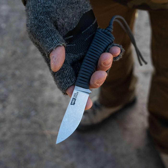 Hand holding the MINI STONED GOAT knife in black, featuring a sleek blade and paracord handle, ideal for outdoor use.