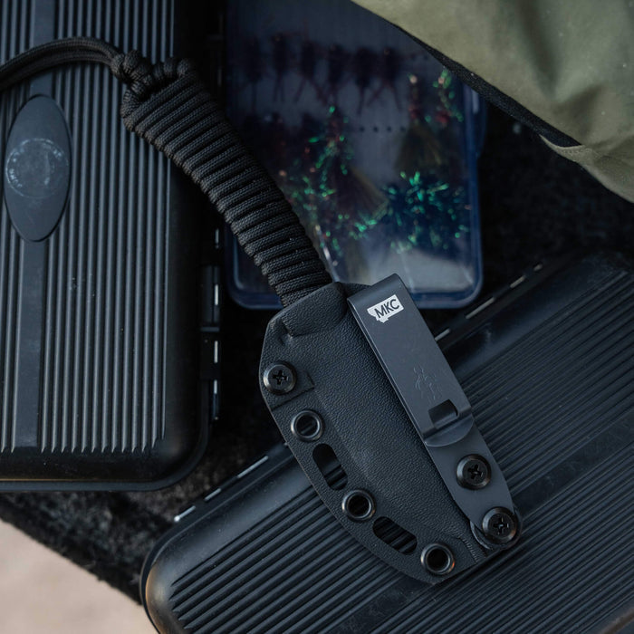 Black MINI STONED GOAT knife with a sheath, resting on tactical gear and fishing tackle boxes.