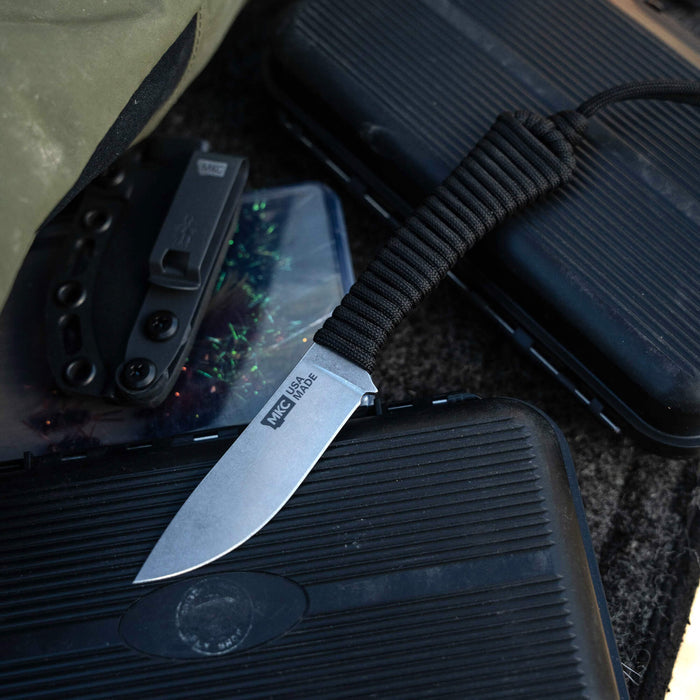 Black MINI STONED GOAT knife resting on textured cases, showcasing its sleek blade and paracord handle.