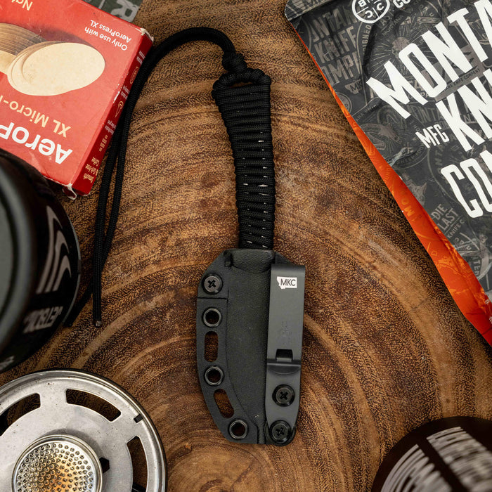 Mini Stoned Goat knife in black with a paracord handle, surrounded by outdoor gear and coffee products on a wooden surface.