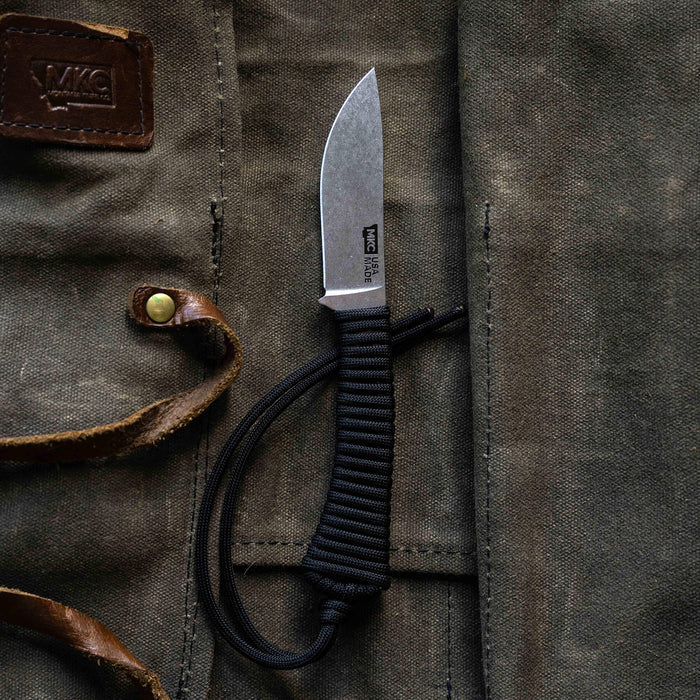 Black MINI STONED GOAT knife with a paracord handle, resting on a rugged canvas background.