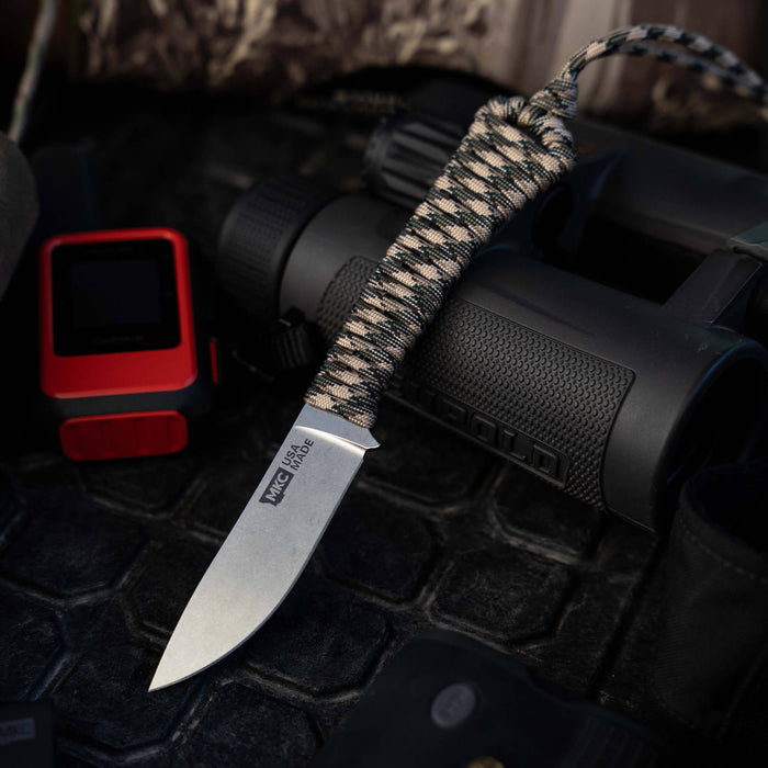 Mini Stoned Goat knife in desert camo with a paracord handle, resting on a textured surface near outdoor gear.