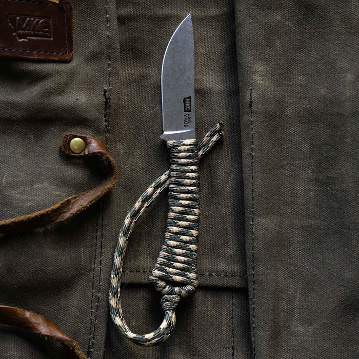 Mini Stoned Goat knife in desert camo with a paracord handle, resting on a textured fabric background.