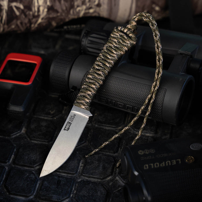 Mini Stoned Goat knife in forest camo with paracord handle, surrounded by outdoor gear and tools. Ideal for hunting and outdoor use.