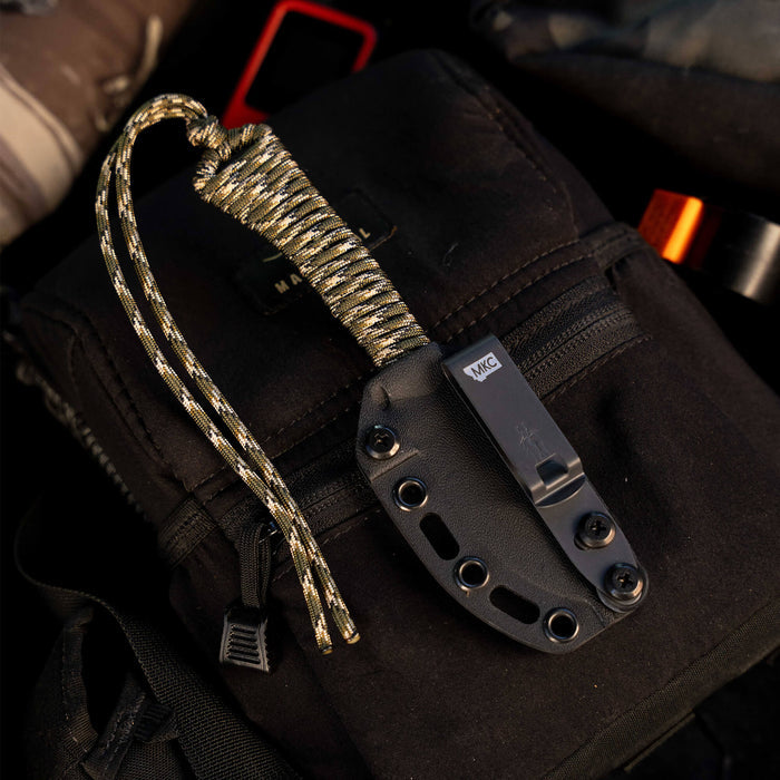 Forest camo MINI STONED GOAT knife with a paracord handle, resting on a black backpack in an outdoor setting.
