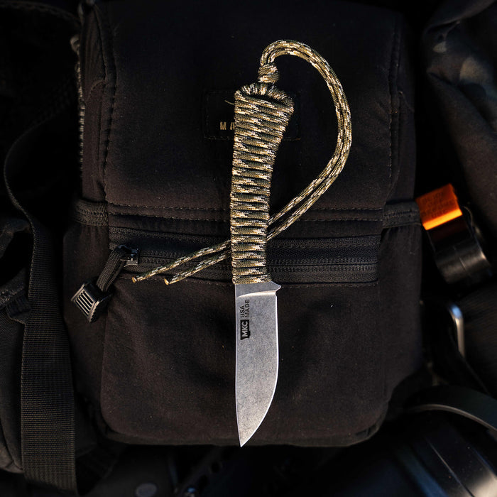 Mini Stoned Goat knife in forest camo with a paracord handle, resting on a black tactical bag. Ideal for outdoor use.