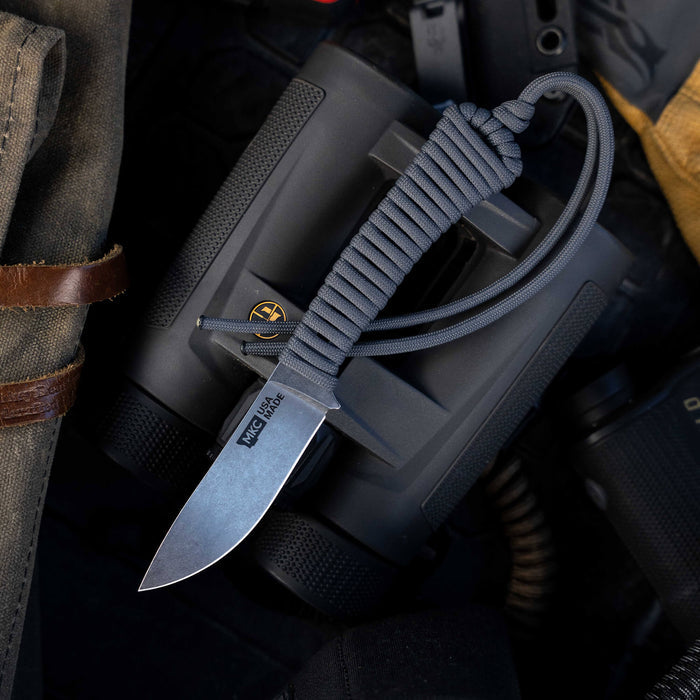 Mini Stoned Goat knife in grey with a paracord handle, resting on a tactical background with gear.
