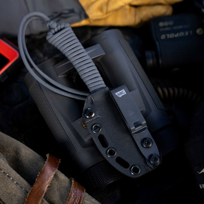 Grey MINI STONED GOAT knife with a paracord handle, secured in a black sheath, resting on outdoor gear.