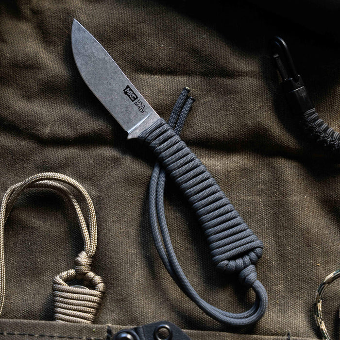 Mini Stoned Goat knife in grey with a paracord handle, resting on a textured fabric background. Ideal for outdoor use.