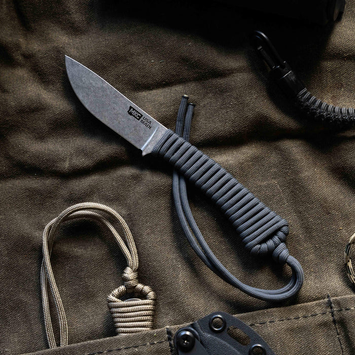 Mini Stoned Goat knife in grey with a paracord handle, resting on a dark fabric background with gear.