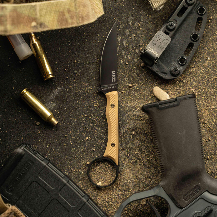 MINI WARGOAT knife with a black blade and tan handle, surrounded by tactical gear and ammunition on a textured surface.