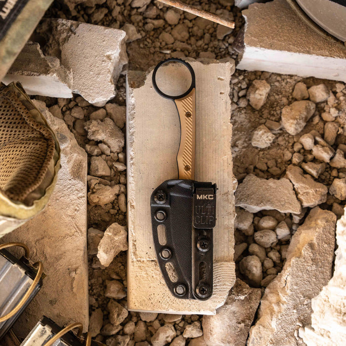 Mini Wargoat knife with black and coyote handle resting on a concrete block among gravel and tools.