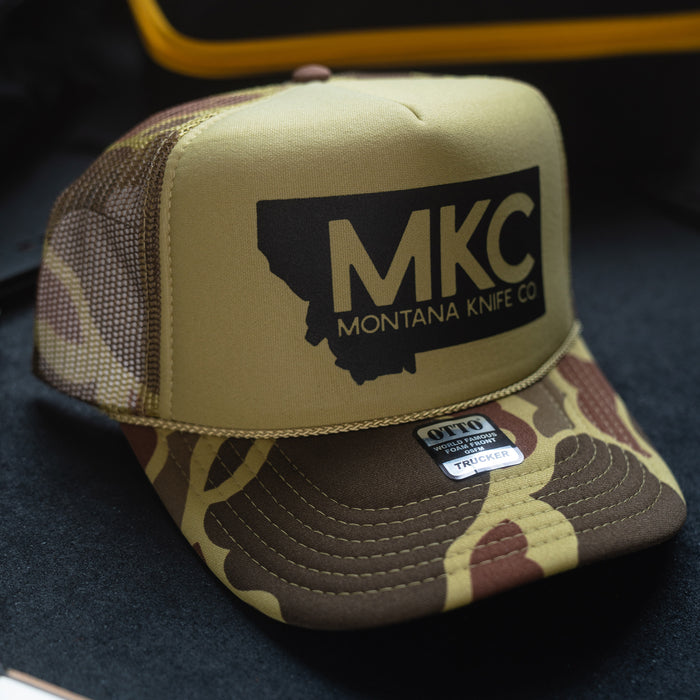 Duck camo foam trucker hat featuring the MKC logo and Montana Knife Company branding, ideal for outdoor enthusiasts.