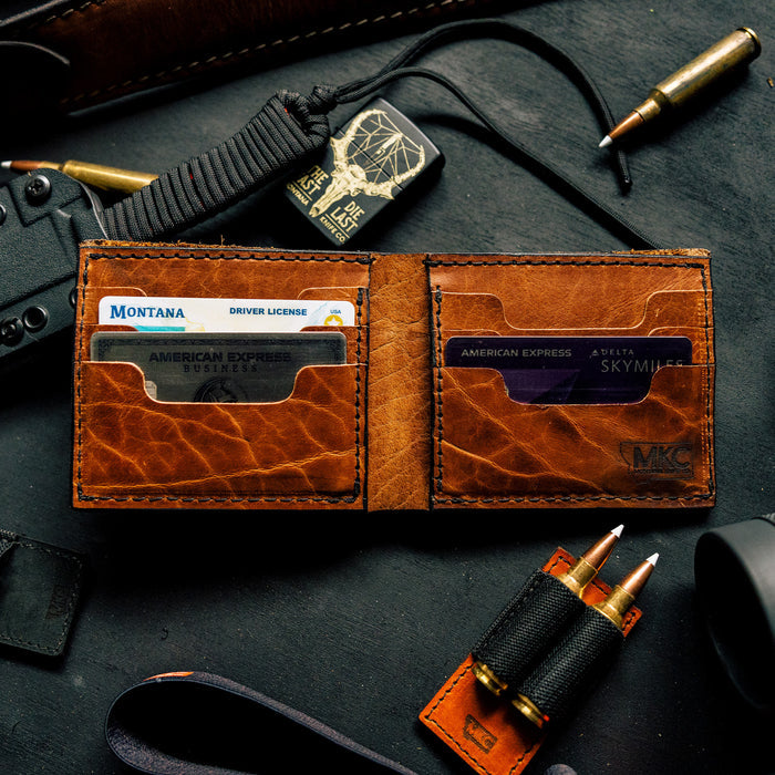 MKC Bison Skull leather wallet open, showcasing card slots and rich brown leather, surrounded by outdoor gear.